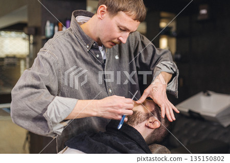 Close up of professional barber shaving male neck with straight razor in barbershop. Beard contouring, classic wet shave, men grooming service and barber work process concept. Close up of professional barber shaving male neck with straight razor in barbershop. Beard contouring, classic wet shave, men grooming service and barber work process concept. 135150802