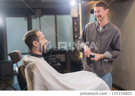 Barber and male client laughing during haircut in barbershop. Friendly atmosphere, professional grooming service, casual conversation, modern men barber salon interior. Barber and male client laughing during haircut in barbershop. Friendly atmosphere, professional grooming service, casual conversation, modern men barber salon interior. 135150803