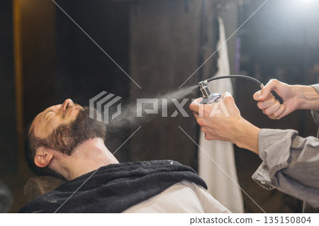 Barber spraying beard and face with water mister during shaving preparation in barbershop. Classic wet shave ritual, men grooming service and professional barber care concept. 135150804