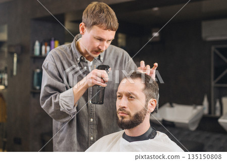 Barber applying hair styling spray to male client during haircut in modern barbershop. Professional grooming service, men hairstyle care, barber work process and lifestyle concept. Barber applying hair styling spray to male client during haircut in modern barbershop. Professional grooming service, men hairstyle care, barber work process and lifestyle concept. 135150808