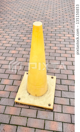 Bright yellow plastic traffic cone standing on a red brick paved sidewalk in a city outdoor area 135150853