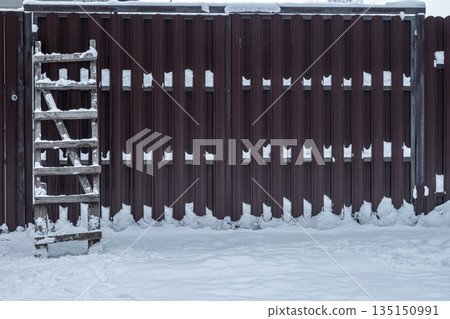 Old wooden ladder leaning against a fence covered with snow. Winter rural background, construction and maintenance concept outdoors. 135150991