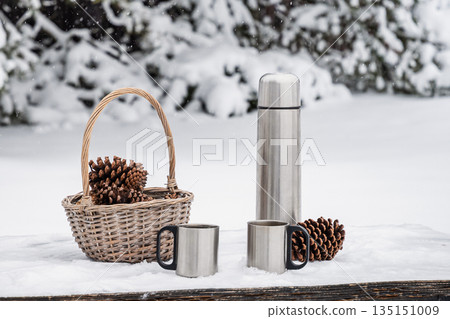 Thermos flask, metal mugs and pinecones on a snow covered wooden table. Winter nature concept, outdoor relaxation, cozy cold season atmosphere. 135151009