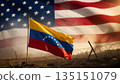 venezuelan flag on a field with barbed wire against a blurred usa flag and bright light in a haze 135151079