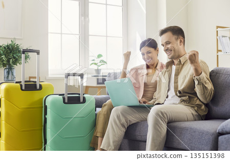 Excited couple booking holiday online at home, cheering with luggage and laptop 135151398