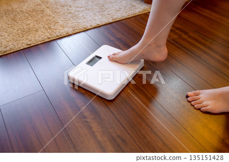 一位年輕女子在客廳裡稱體重。她的腳放在秤上。她身材苗條,膚色呈橘色。 一位年輕女子在客廳裡稱體重。她的腳放在秤上。她身材苗條,膚色呈橘色。 135151428