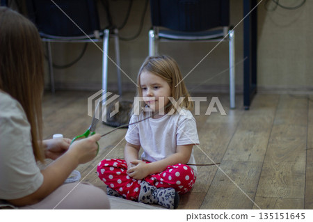 A mother and a small child sit on the floor, making a craft out of a stick with scissors. The family spends time being creative at home, encouraging the child's early development and imagination. A mother and a small child sit on the floor, making a craft out of a stick with scissors. The family spends time being creative at home, encouraging the child's early development and imagination. 135151645