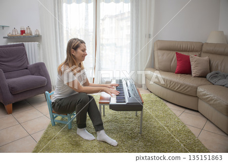 Woman sitting barefoot on a small chair in a bright living room, playing an electronic keyboard on a low table, practicing and enjoying music as a relaxing home hobby and learning activity Woman sitting barefoot on a small chair in a bright living room, playing an electronic keyboard on a low table, practicing and enjoying music as a relaxing home hobby and learning activity 135151863