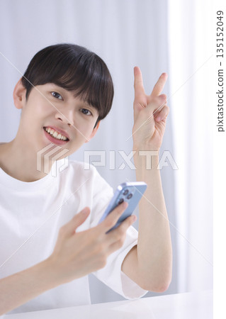 A young man making a V sign with his smartphone in his living room A young man making a V sign with his smartphone in his living room 135152049