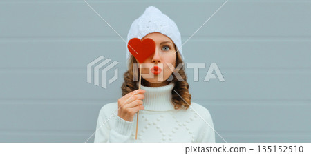 Winter christmas portrait of beautiful happy young woman blowing kiss with lollipop in white hat 135152510