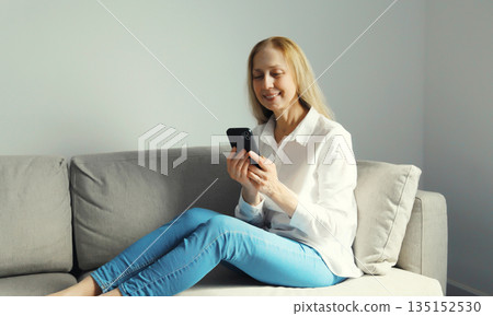 Happy mature woman looks at phone, girl with smartphone sitting on the couch in room at home Happy mature woman looks at phone, girl with smartphone sitting on the couch in room at home 135152530