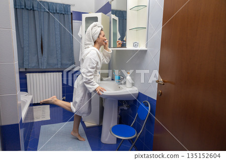 Woman standing in a bathroom wearing a bathrobe and towel wrapped around her head, brushing her teeth in front of a mirror with a confident and healthy expression, making a playful pose Woman standing in a bathroom wearing a bathrobe and towel wrapped around her head, brushing her teeth in front of a mirror with a confident and healthy expression, making a playful pose 135152604