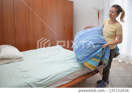 Woman in yellow tee and pajamas smoothing a white duvet over a bed, tidying sheets and pillows as part of a morning routine in a cozy, orderly bedroom setting Woman in yellow tee and pajamas smoothing a white duvet over a bed, tidying sheets and pillows as part of a morning routine in a cozy, orderly bedroom setting 135152636