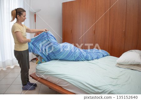 Woman smoothing a blue striped duvet on a neatly made bed in a bright bedroom, carrying out morning housekeeping and tidying as part of a daily domestic routine and self care ritual Woman smoothing a blue striped duvet on a neatly made bed in a bright bedroom, carrying out morning housekeeping and tidying as part of a daily domestic routine and self care ritual 135152640