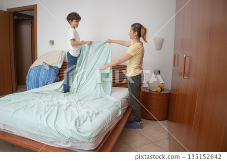 Mother and son making the bed together in a bright bedroom, teaching responsibility and teamwork while sharing domestic chores, smiling and bonding during a cozy morning routine 135152642