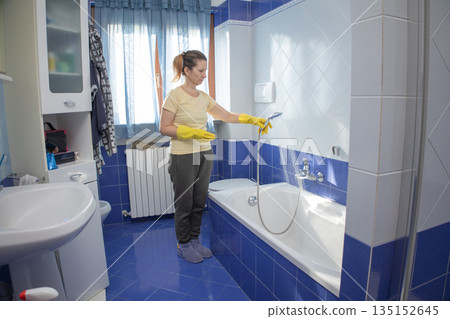 Woman wearing yellow protective gloves and holding a showerhead, rinsing the bathtub with water while doing household chores in a clean, tiled bathroom Woman wearing yellow protective gloves and holding a showerhead, rinsing the bathtub with water while doing household chores in a clean, tiled bathroom 135152645