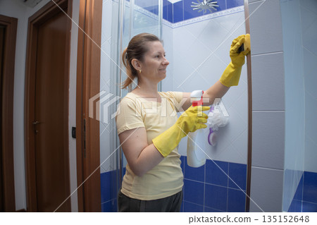 Woman wearing yellow rubber gloves and holding a spray bottle, diligently cleaning bathroom shower tiles to maintain a hygienic and sparkling home environment 135152648