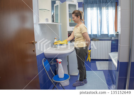 Woman wearing rubber gloves and slippers, diligently cleaning a bathroom sink with a sponge and cleaner, maintaining hygiene and order in a domestic setting 135152649