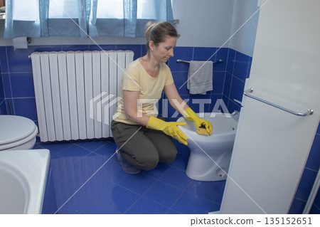Woman in yellow rubber gloves scrubbing a bidet with a sponge, cleaning bathroom surfaces for hygiene and home maintenance as part of everyday household chores and care 135152651