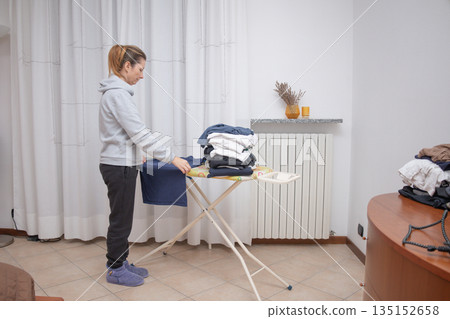 Woman folding clean clothes and organizing a pile of ironed garments on an ironing board inside a bright room at home, performing daily domestic chores Woman folding clean clothes and organizing a pile of ironed garments on an ironing board inside a bright room at home, performing daily domestic chores 135152658