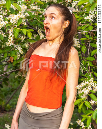 Young woman sneezes in the park against the background of a flowering tree. Allergy to pollen concept. 135153231