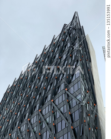 Upward view of a modern high-rise with dark glass windows, diagonal black steel bracing and small orange details against grey sky. Upward view of a modern high-rise with dark glass windows, diagonal black steel bracing and small orange details against grey sky. 135153991