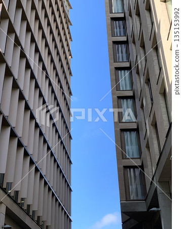 LONDON, UK, Low angle view between two modern buildings with geometric facade and windows against clear blue sky LONDON, UK, Low angle view between two modern buildings with geometric facade and windows against clear blue sky 135153992