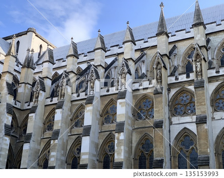 London close view of Westminster Abbey side facade with Gothic buttresses, statues, pointed arches and stained glass windows under blue sky. London close view of Westminster Abbey side facade with Gothic buttresses, statues, pointed arches and stained glass windows under blue sky. 135153993