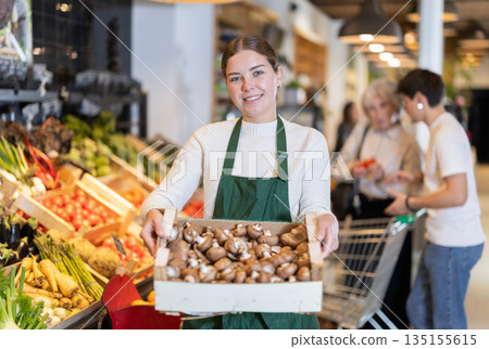 Girl employee in dark green apron stands in sales area with box of mushrooms Girl employee in dark green apron stands in sales area with box of mushrooms 135155615