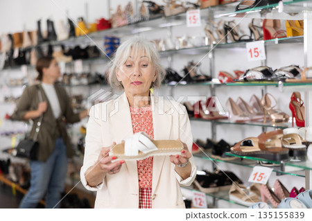 Next to shelf of womens shoes,mature lady picks up stable,comfortable pair of shoes 135155839