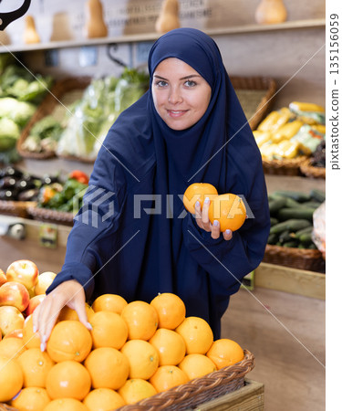 Smiling european woman in muslim dress selecting oranges in supermarket 135156059