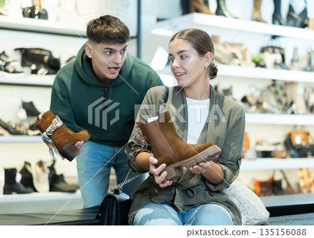 Young guy and girl choosing demi-season womens boots in shoe store Young guy and girl choosing demi-season womens boots in shoe store 135156088