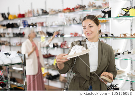 Positive young female customer holds in hands and compares slippers Positive young female customer holds in hands and compares slippers 135156098