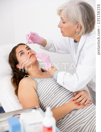 Mature woman beautician performing procedure of injection biorevitalization to young woman for alignment of facial asymmetry in esthetic clinic 135156158