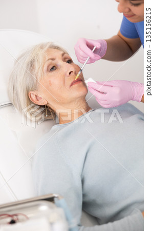 Young woman cosmetologist in uniform making injections for lip augmentation and anti-wrinkle of elderly female in esthetic clinic indoors 135156215