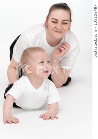 Playful little boy of one year old crawling on all fours on floor and happily looks ahead. Caucasian mommy is behind baby child and watches son play. Female parent and toddler wearing white t-shirts Playful little boy of one year old crawling on all fours on floor and happily looks ahead. Caucasian mommy is behind baby child and watches son play. Female parent and toddler wearing white t-shirts 135158497