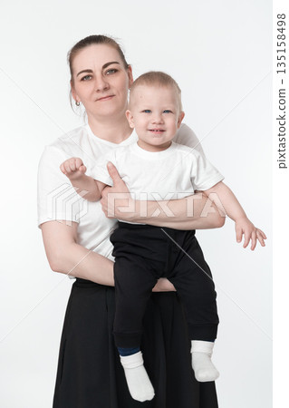 Middle aged Caucasian mother holding 1 year old boy's child in her arms and hugging darling son. Mommy and toddler boy looking at camera. Female parent and kid wearing white t-shirts and black bottoms Middle aged Caucasian mother holding 1 year old boy's child in her arms and hugging darling son. Mommy and toddler boy looking at camera. Female parent and kid wearing white t-shirts and black bottoms 135158498