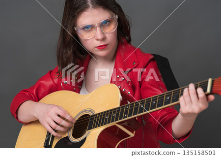 Close-up view of female guitarist strumming strings of her classical acoustic guitar while looking directly at camera. Stylish musician is young Caucasian woman wearing glasses and red leather jacket 135158501