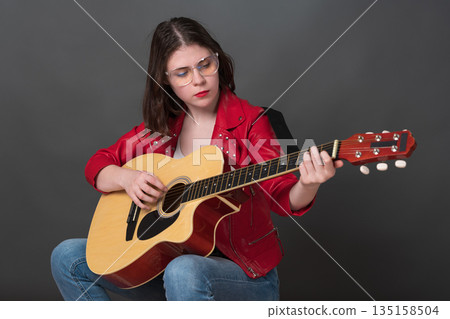 Nice woman is practicing her acoustic guitar and creating warm and lively atmosphere. Young adult female musician, wearing glasses and vibrant red leather jacket, adds to the performance's vitality 135158504
