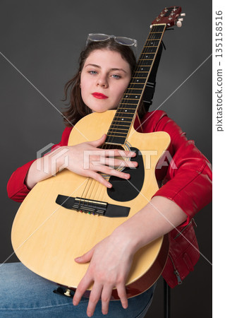 Female guitarist with Spanish six string acoustic guitar gently nestles musical instrument against her chest as she looks at camera. She is young female musician wearing red leather jacket Female guitarist with Spanish six string acoustic guitar gently nestles musical instrument against her chest as she looks at camera. She is young female musician wearing red leather jacket 135158516