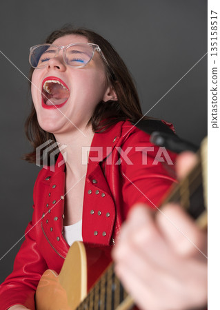 Woman in euphoria plays guitar and sings song with great fervor. Rock musician in state of pure bliss, with her eyes closed, mouth wide open, and voice resonating with a powerful emotional intensity 135158517