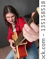Woman playing guitar in lighthearted manner, appears to be deriving enjoyment from activity. She is doing so with smiling. Young female exudes passion and is attired in crimson leather jacket, glasses Woman playing guitar in lighthearted manner, appears to be deriving enjoyment from activity. She is doing so with smiling. Young female exudes passion and is attired in crimson leather jacket, glasses 135158518