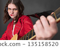 Playful woman guitarist is enjoying herself is playing guitar and she is doing so with her lips pursed. Passionate young female wearing eyeglasses, is dressed crimson leather jacket Playful woman guitarist is enjoying herself is playing guitar and she is doing so with her lips pursed. Passionate young female wearing eyeglasses, is dressed crimson leather jacket 135158520