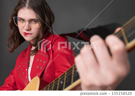 Playful woman guitarist is enjoying herself is playing guitar and she is doing so with her lips pursed. Passionate young female wearing eyeglasses, is dressed crimson leather jacket 135158520