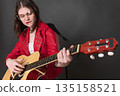 Playful woman is strumming an acoustic guitar with her lips pursed and looks at strings while playing. Passionate young woman wearing eyeglasses. 18 year old musician is dressed in red leather jacket Playful woman is strumming an acoustic guitar with her lips pursed and looks at strings while playing. Passionate young woman wearing eyeglasses. 18 year old musician is dressed in red leather jacket 135158521