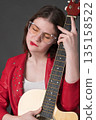 Lady with guitar gently nestles musical instrument against her bosom. A young female musician wearing glasses and red leather jacket Lady with guitar gently nestles musical instrument against her bosom. A young female musician wearing glasses and red leather jacket 135158522