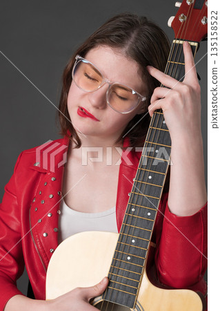 Lady with guitar gently nestles musical instrument against her bosom. A young female musician wearing glasses and red leather jacket 135158522