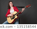 Confident young woman guitarist strumming acoustic guitar and looking down at strings with concentration. Stylish woman musician wearing eyeglasses, dressed crimson leather jacket and blue jeans Confident young woman guitarist strumming acoustic guitar and looking down at strings with concentration. Stylish woman musician wearing eyeglasses, dressed crimson leather jacket and blue jeans 135158544