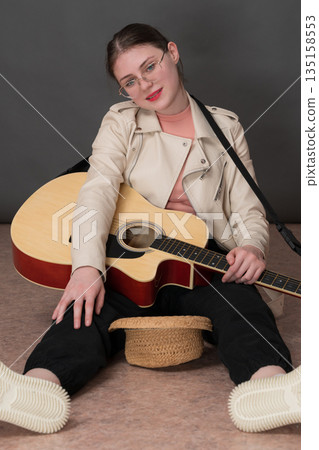 Street musician with guitar sitting with legs spread and straw hat in front of her, also intended as donation for playing music. Her face pointed at camera, conveying feeling of hope and anticipation 135158553