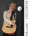 Young female hipster musician holding Spanish six string acoustic guitar in front of her hugging string instrument with both hands and looking at camera. Woman in beige leather jacket and straw hat Young female hipster musician holding Spanish six string acoustic guitar in front of her hugging string instrument with both hands and looking at camera. Woman in beige leather jacket and straw hat 135158556